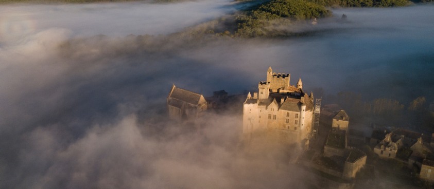 Vue aérienne d’un château médiéval émergeant de la brume, entouré de collines boisées au lever du jour